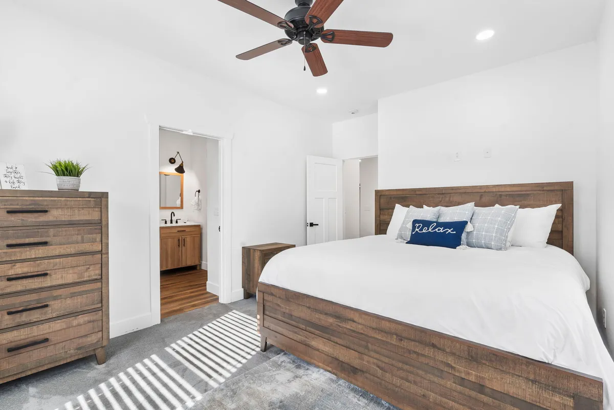 A serene queen bedroom with a wooden bed frame adorned with decorative pillows, a wooden dresser, and an open door leading into a connected bathroom. The natural light and calming colors create a comfortable and welcoming environment.