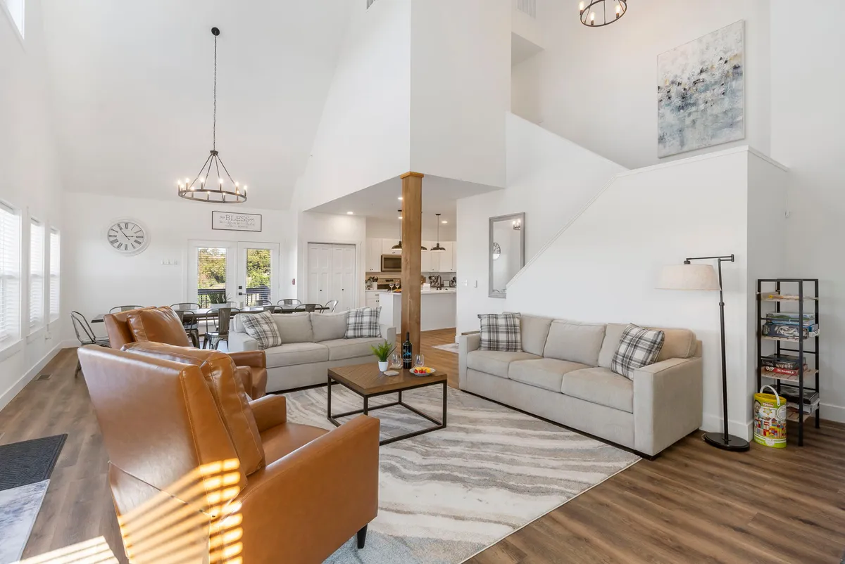 The image showcases a beautifully decorated living room with plush sofas arranged around a stylish coffee table, an elegant chandelier overhead, and a striking staircase in the background. The room exudes warmth and comfort, inviting guests to gather for game nights, cozy conversations, or peaceful reading, all in a light-filled, high-end space.