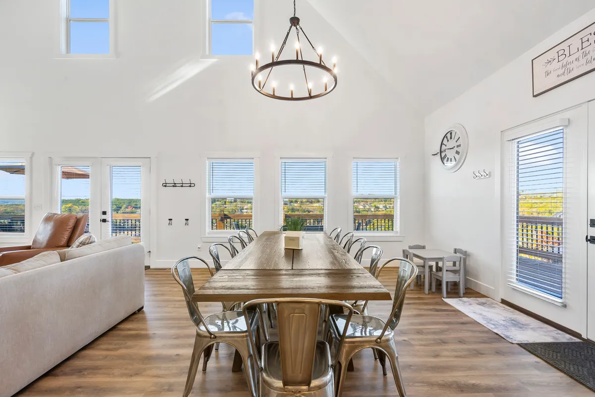 This interior image showcases a long wooden dining table beneath a chandelier, surrounded by a comfortable seating area and large windows that offer natural light and views. The setup reflects an inviting atmosphere perfect for family dining, celebrations, and social gatherings.