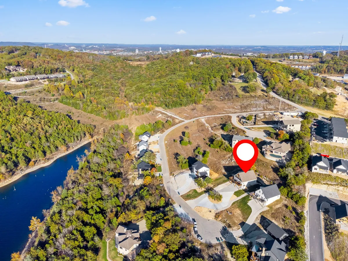 A tranquil exterior view featuring several houses nestled near a shimmering lake, surrounded by lush greenery and a gently curving road. The image captures the peaceful community setting and hints at the stunning sunset possibilities over the water, with the natural beauty of the Ozarks on full display.