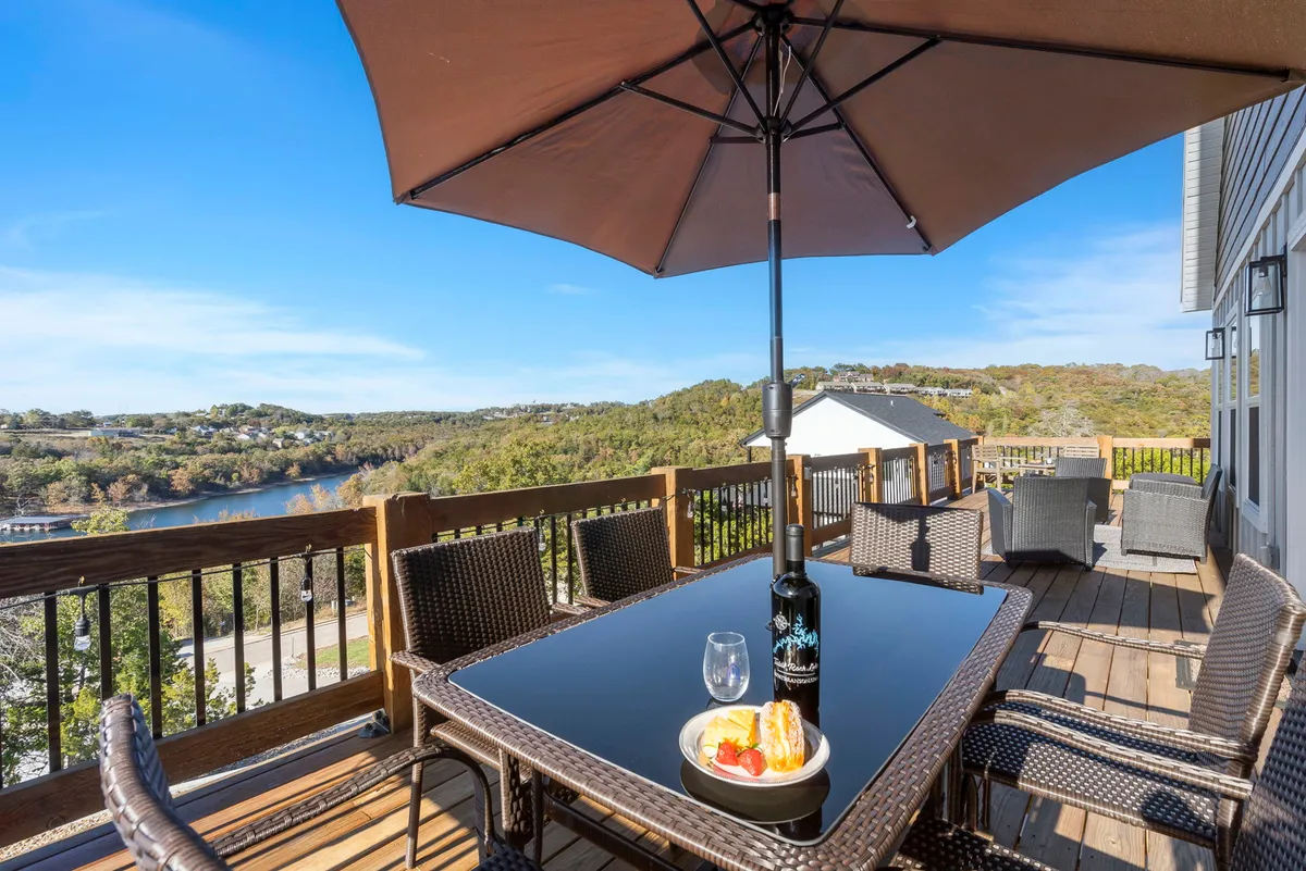 An outdoor dining table sits under a large umbrella, set with food and drinks, overlooking a scenic landscape of rolling hills. The inviting setup is perfect for social gatherings and relaxed evenings with friends, capturing the essence of leisurely group enjoyment and a connection to nature.