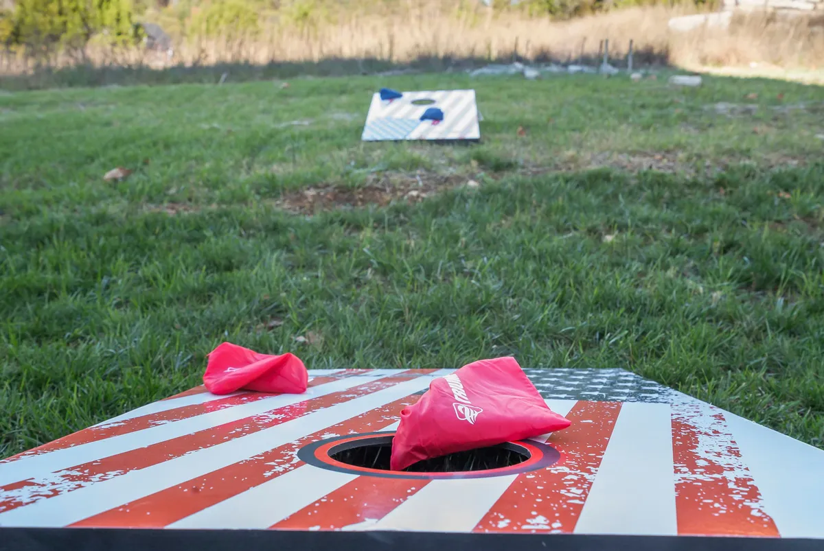 A playful outdoor scene featuring cornhole boards and bright red bags set up on the grass, inviting guests to enjoy friendly games and social picnics in a casual, family-friendly atmosphere.