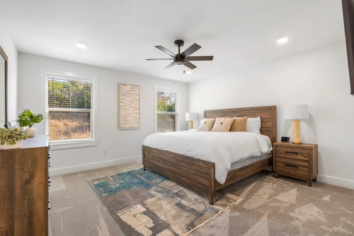 A tranquil king-sized bedroom featuring large windows that flood the room with natural light, wooden nightstands on either side, and decorative wall art above the bed. The bedding looks plush and inviting, contributing to a serene ambiance ideal for rest and relaxation.