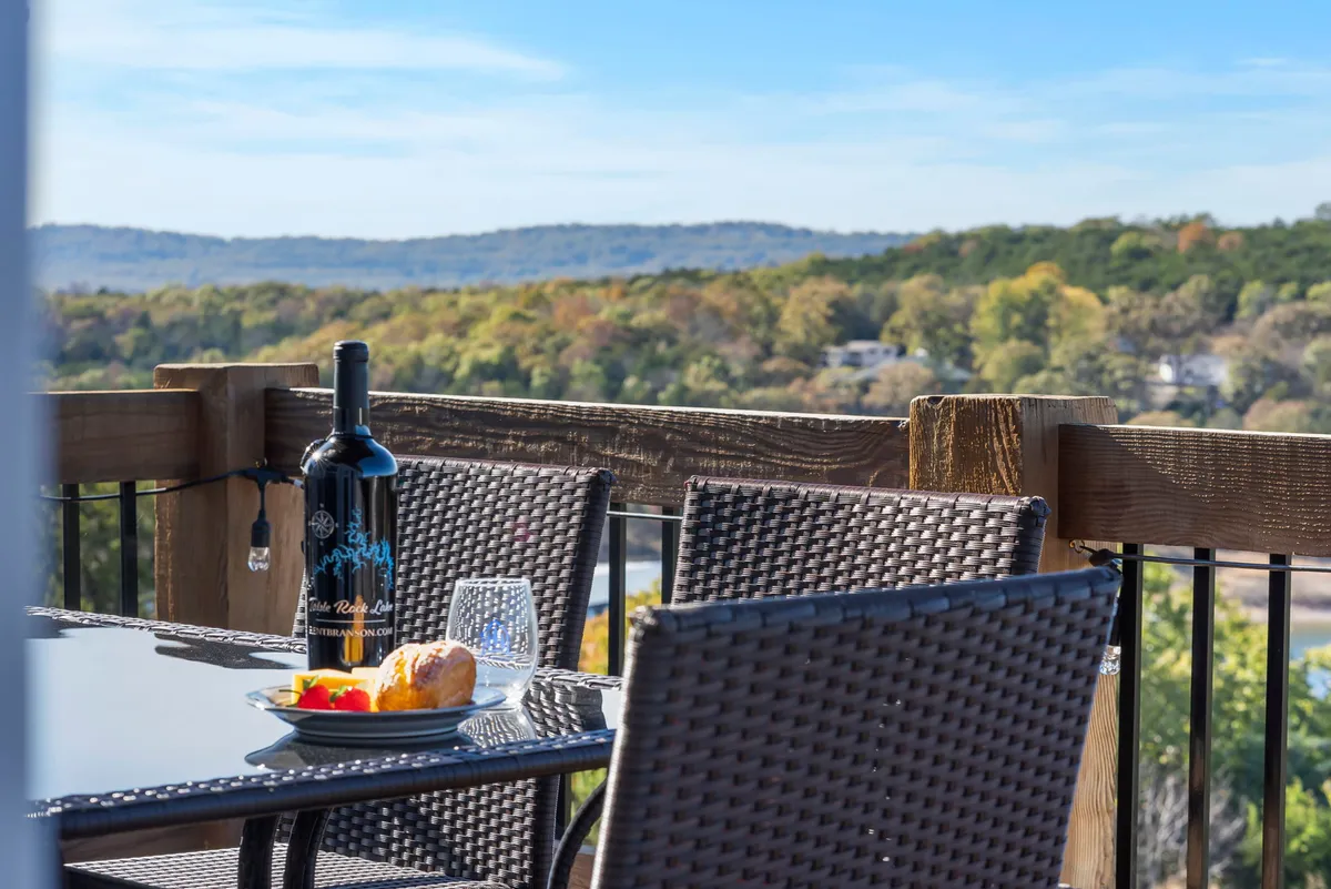 An inviting outdoor dining setup on a deck or patio with a wine bottle, glasses, and a plate of food, overlooking a beautiful natural landscape. The mood is relaxed and luxurious, perfect for enjoying meals or drinks as a group while taking in the scenery.