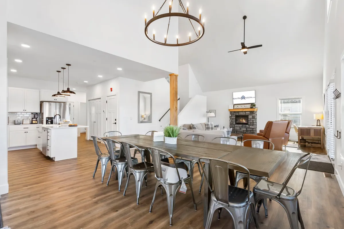 A spacious and inviting dining area inside the home, featuring a long table set for gatherings, a glowing chandelier, a welcoming fireplace, and plush seating. The room radiates warmth, ideal for group meals, shared stories, and cozy evenings.