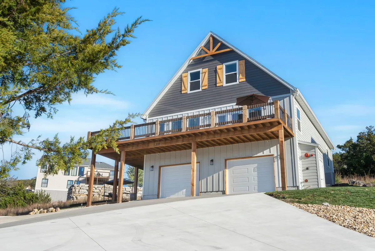 The home’s large deck overlooks a sloping driveway, framed by wooden accents and garage doors. The deck is set up for gatherings, with space to enjoy sunsets and the surrounding landscape—emphasizing shared outdoor moments and the unique hillside location.