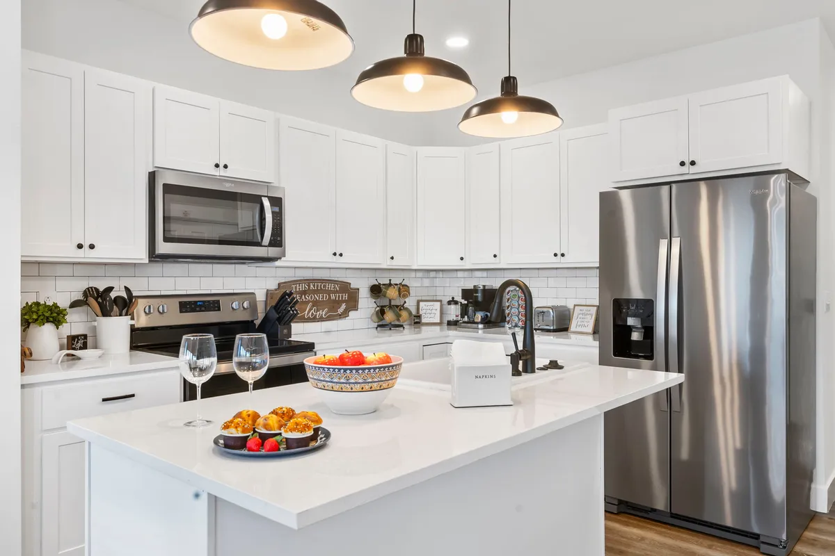 The gourmet kitchen is highlighted in this image, with a spacious island countertop, modern appliances, hanging light fixtures, and decorative touches like a bowl of fruit and wine glasses. The room is designed for cooking, entertaining, and casual gatherings.