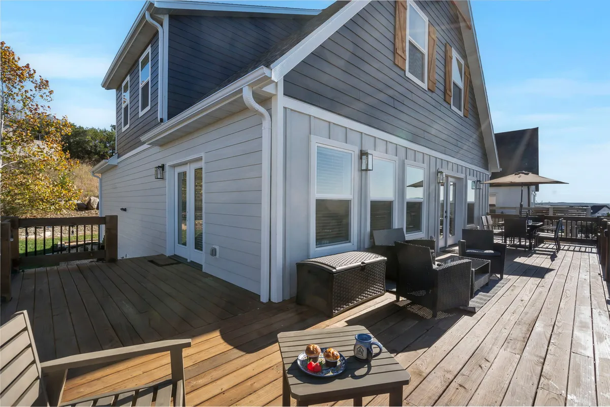 An exterior shot showcasing a spacious deck furnished with comfortable seating and a dining area, surrounded by landscaped greenery. The view extends to a horizon where the sun is rising or setting, creating a tranquil and inviting atmosphere ideal for gatherings.